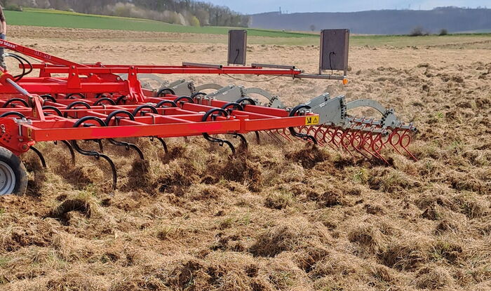 Auf dem Bild ist auf einem Feld eine rote Bodenbearbeitungsmaschine mit schwarzen Federzinken zu erkennen.